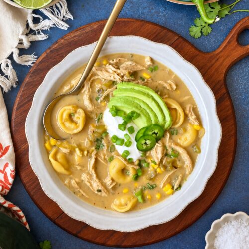 A bowl of creamy soup with tortellini, chicken, corn, sliced avocado, jalapeño, green onions, and a dollop of sour cream, served on a wooden board with limes and peppers nearby.