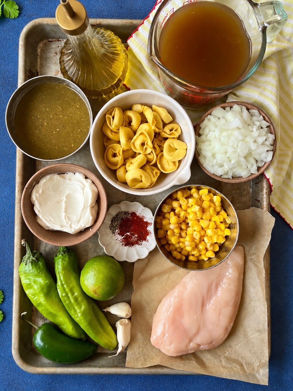 A tray with chicken breast, corn, cheese tortellini, diced onions, green chiles, lime, garlic, broth, salsa verde, cream cheese, olive oil, and spices, arranged in bowls and on parchment.