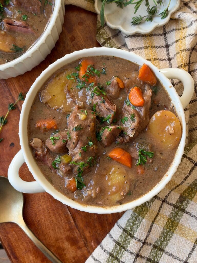 A bowl of beef stew with chunks of beef, potatoes, carrots, and herbs in a brown broth sits on a wooden table next to a spoon and checkered cloth.