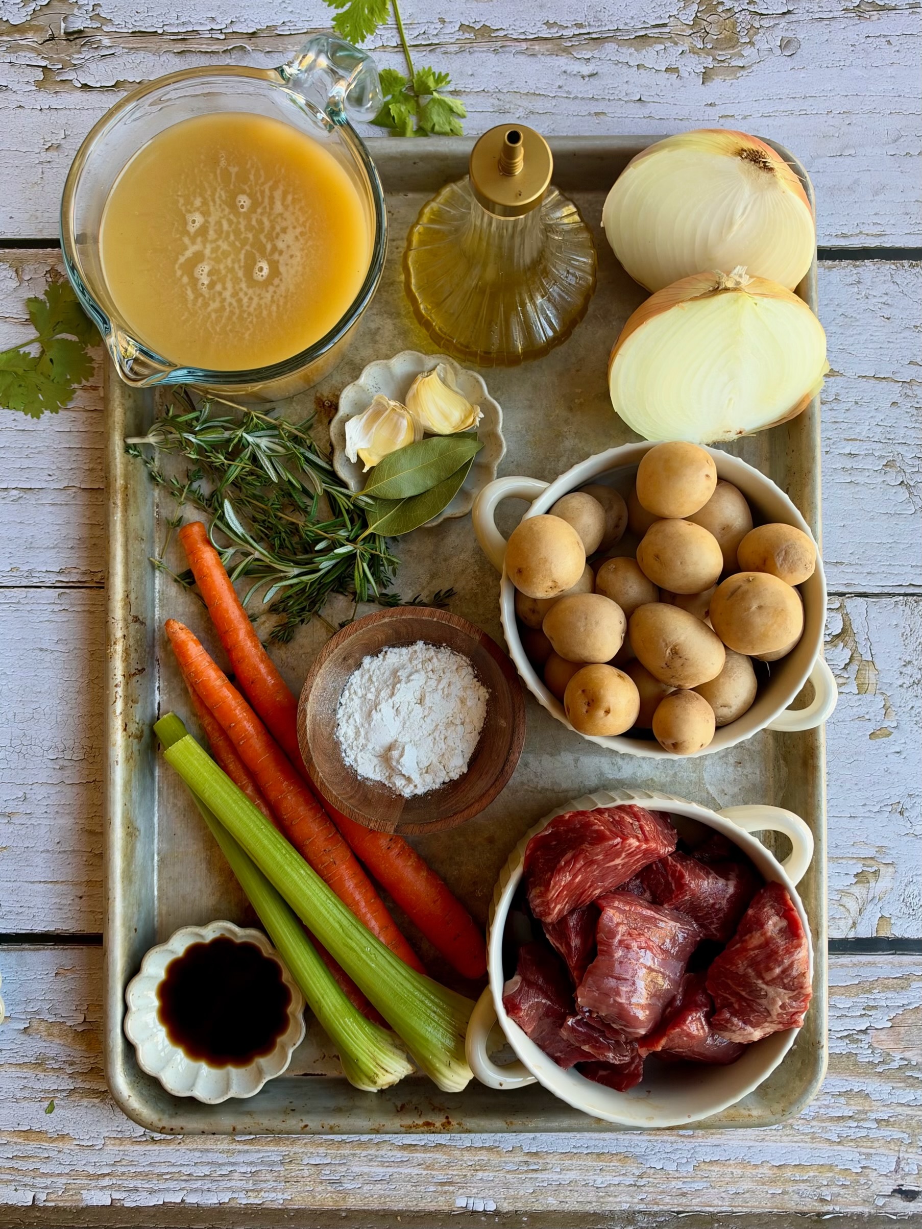 A tray with beef chunks, baby potatoes, carrots, celery, onions, garlic, broth, olive oil, flour, fresh herbs, bay leaves, and soy sauce arranged neatly on a rustic wooden surface.