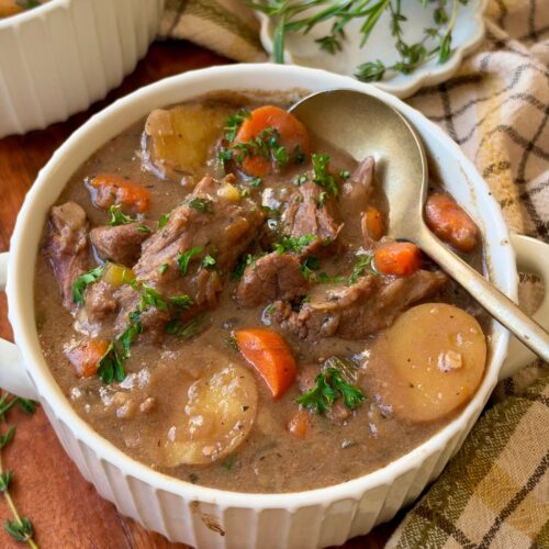 A bowl of beef stew with chunks of beef, carrots, and potatoes, garnished with parsley, with a spoon and fresh herbs on a plaid cloth.