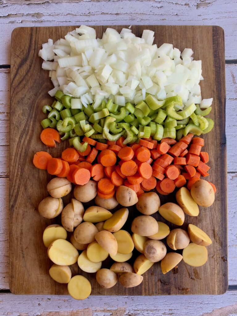 Chopped onions, celery, carrots, and halved baby potatoes are neatly arranged in rows on a wooden cutting board on a light rustic wooden table.