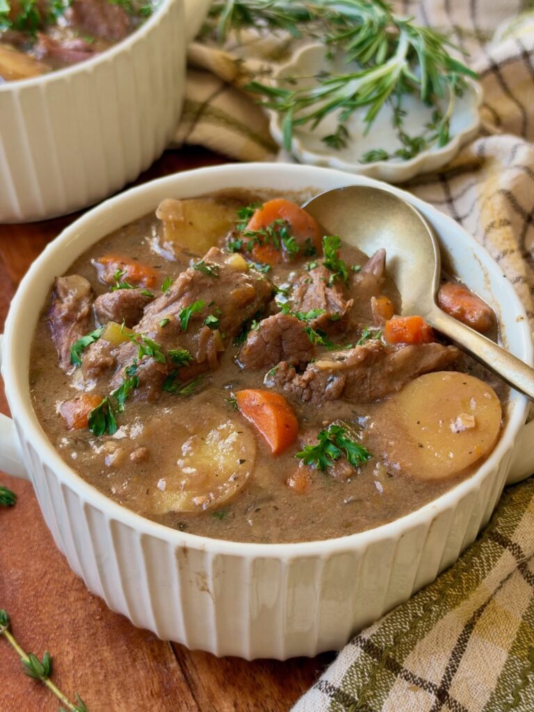 A bowl of hearty beef stew with chunks of beef, potatoes, carrots, and herbs, served with a spoon and garnished with fresh parsley on a wooden table with a plaid cloth and fresh rosemary nearby.
