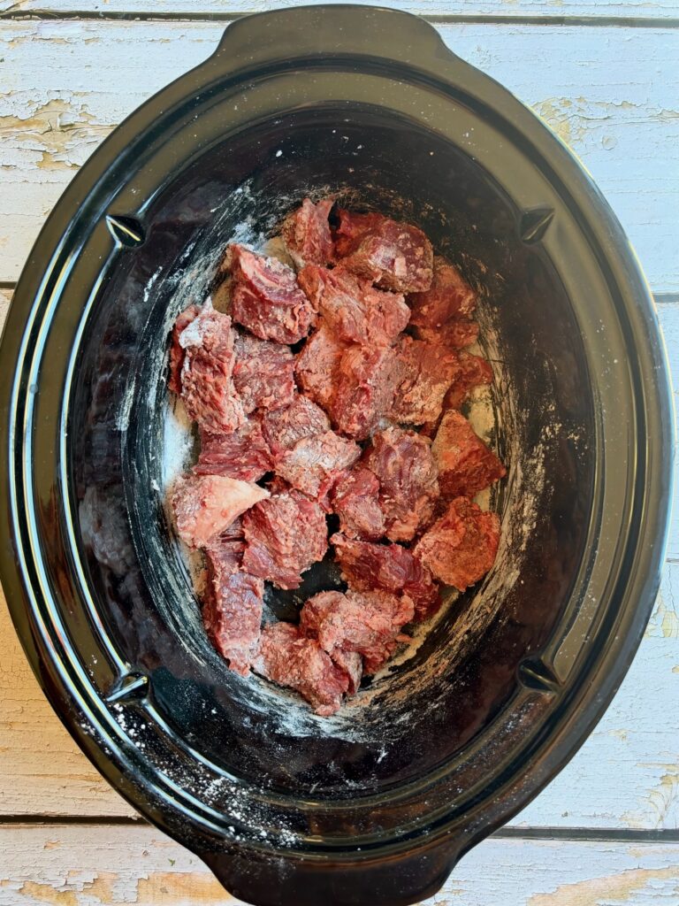 Chunks of raw beef coated with flour are placed in a black slow cooker, sitting on a light wooden surface.