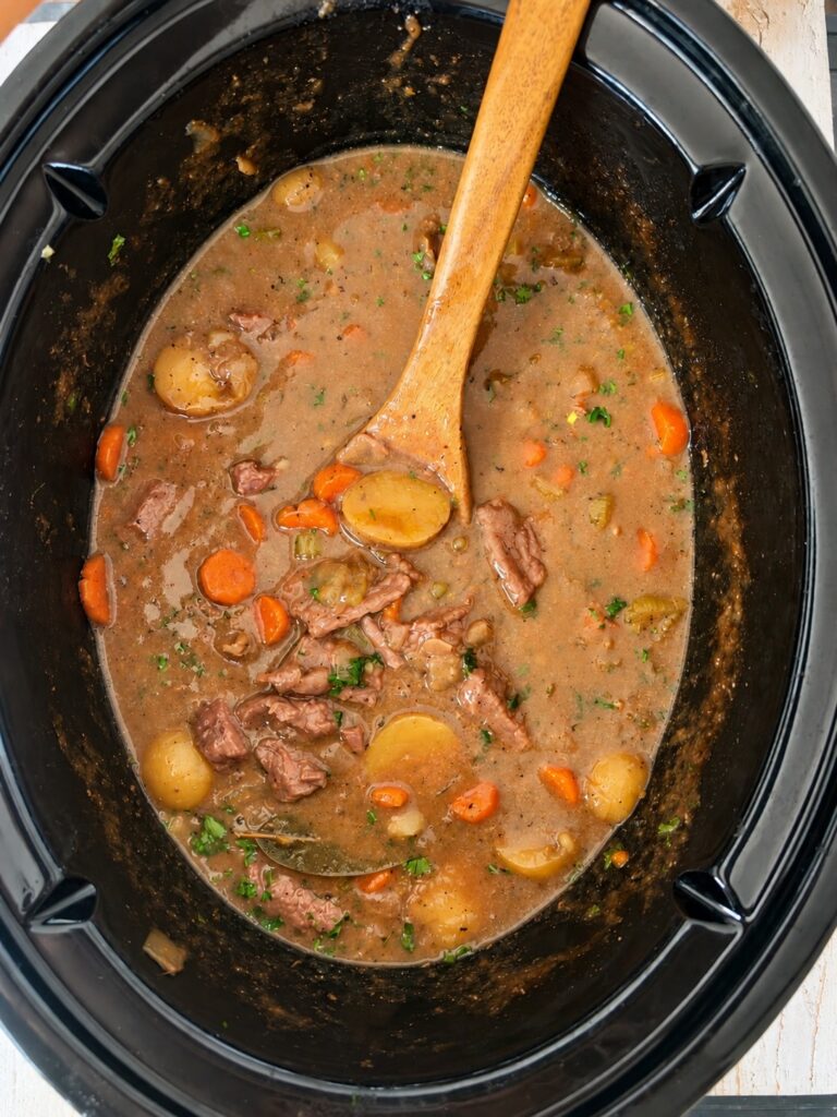 Beef stew with carrots, potatoes, and herbs in a slow cooker. A wooden spoon rests in the stew, mixing the chunky vegetables and meat in a rich brown broth.