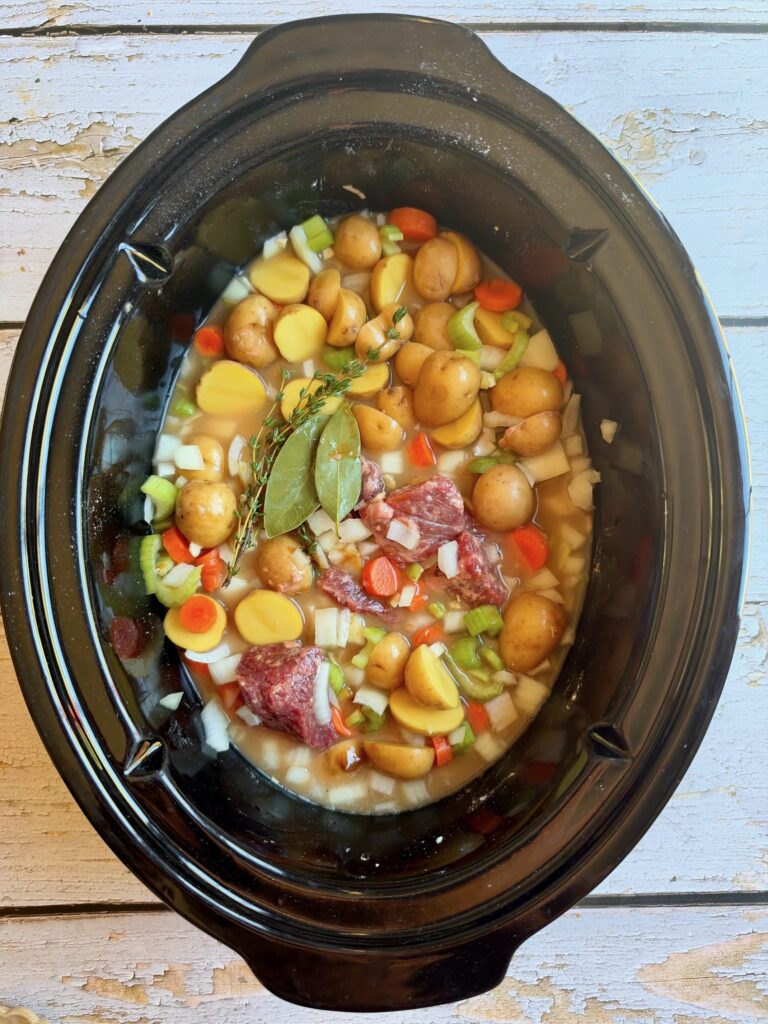 A slow cooker filled with raw stew ingredients: small potatoes, carrots, celery, onions, beef chunks, herbs, and broth, ready to cook.