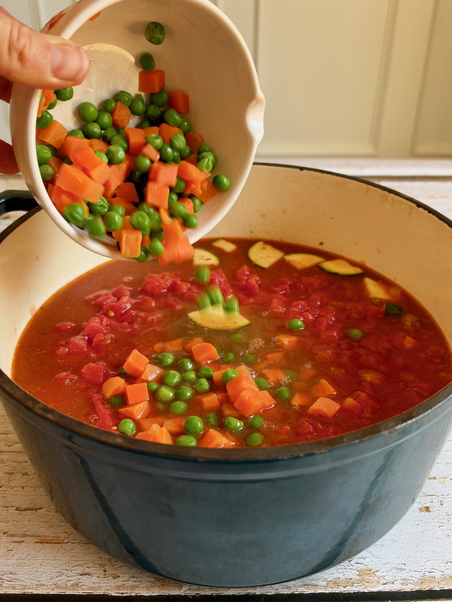 Frozen carrots and peas being added to the beef and vegetable soup in a Dutch oven.