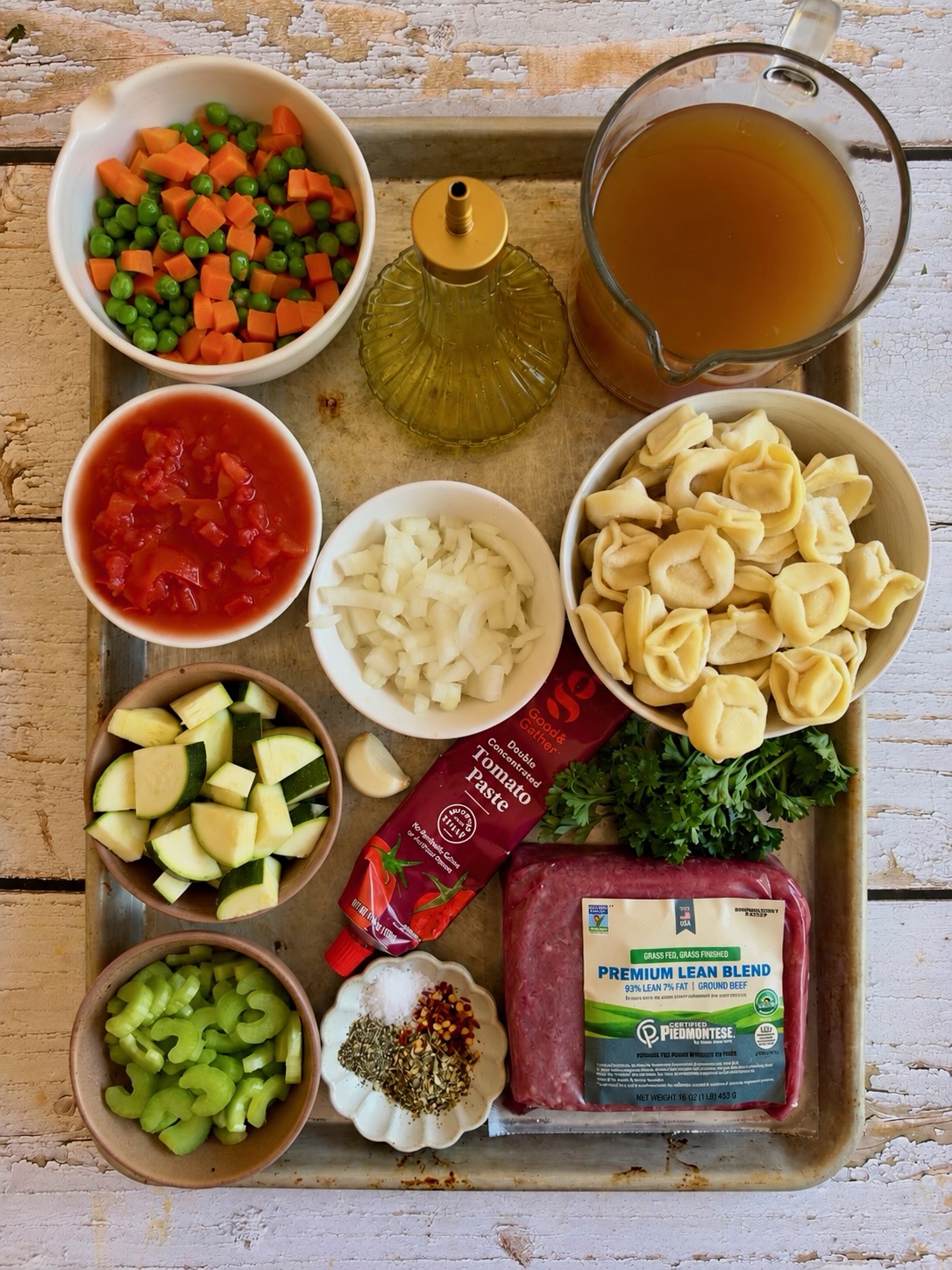 Ingredients for beef and tortellini vegetable soup on a sheet pan, including ground beef, tortellini, diced tomatoes, zucchini, celery, onion, carrots and peas, broth, tomato paste, and spices.