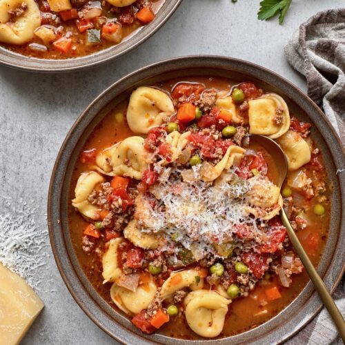 Beef and tortellini vegetable soup topped with grated Parmesan in a dark bowl.