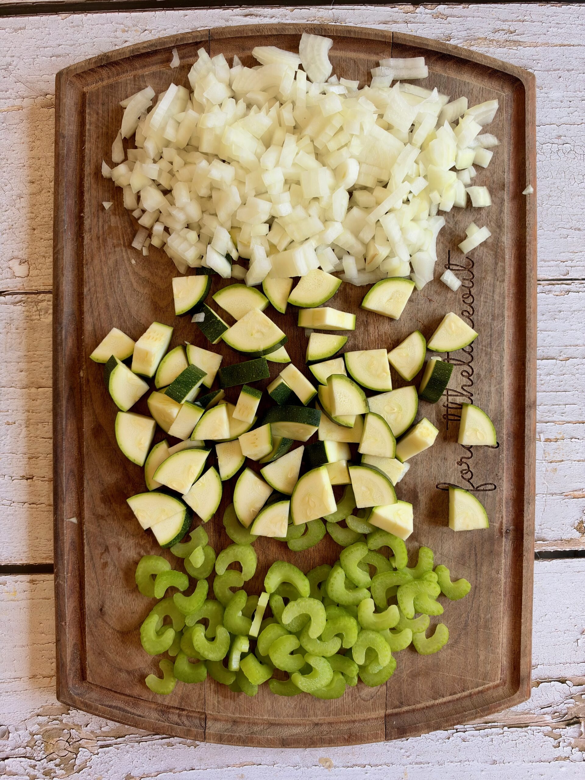 Diced onion, zucchini, and sliced celery on a wooden cutting board.