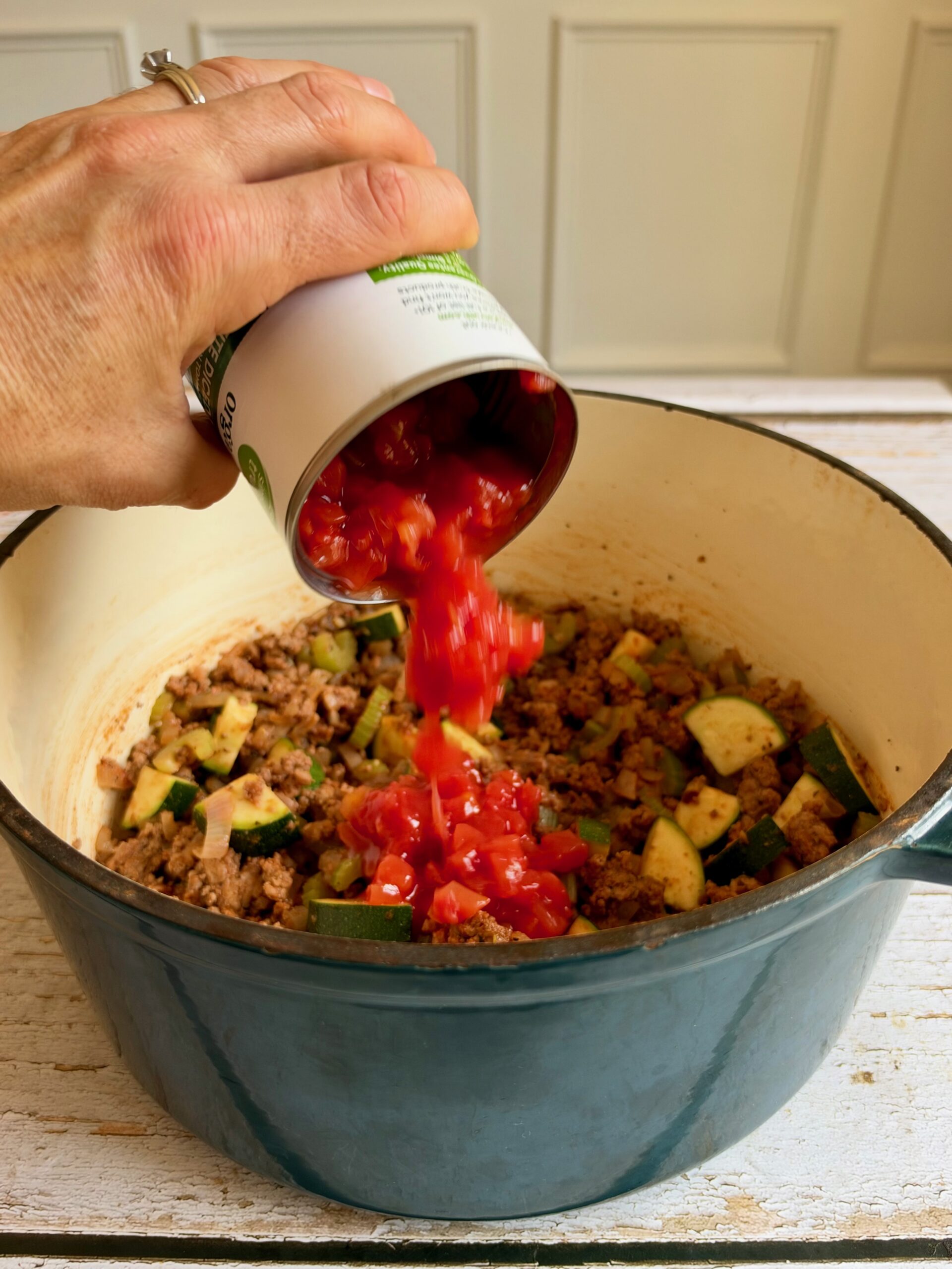 A can of diced tomatoes being poured into ground beef and zucchini in a Dutch oven.