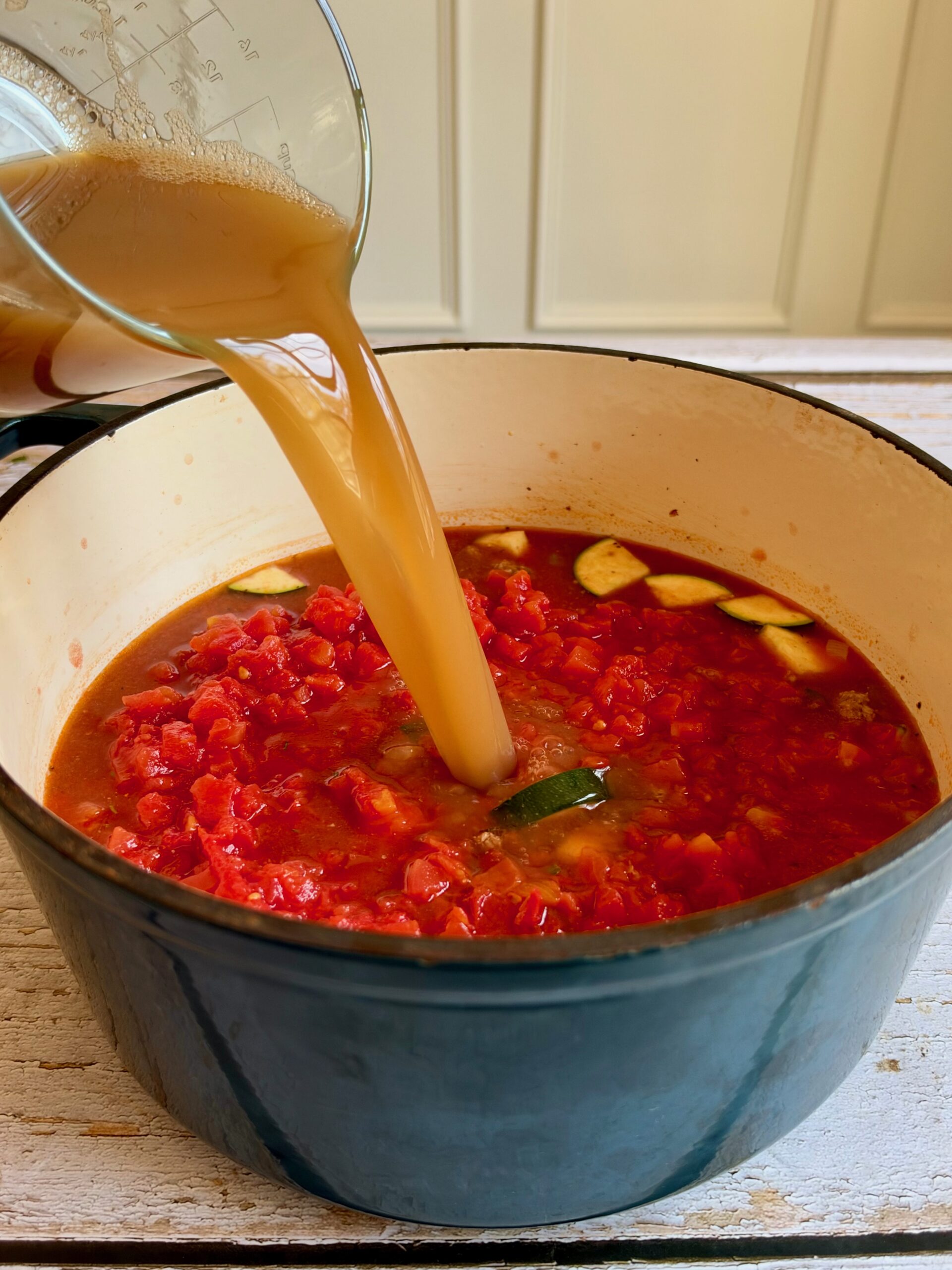 Bone broth being poured into the soup with diced tomatoes in a Dutch oven.