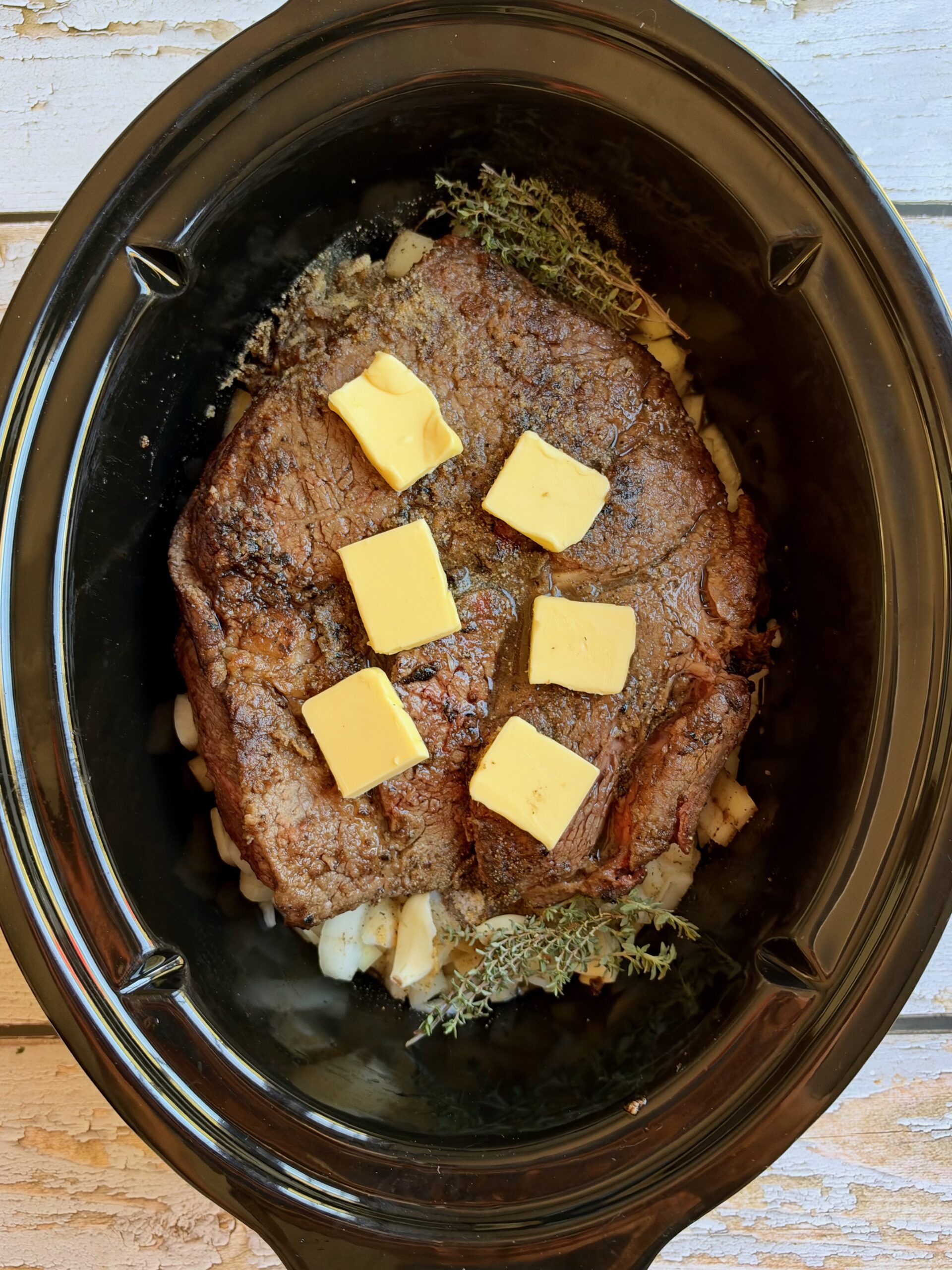 Seared chuck roast topped with butter slices and fresh thyme in a slow cooker over onions.