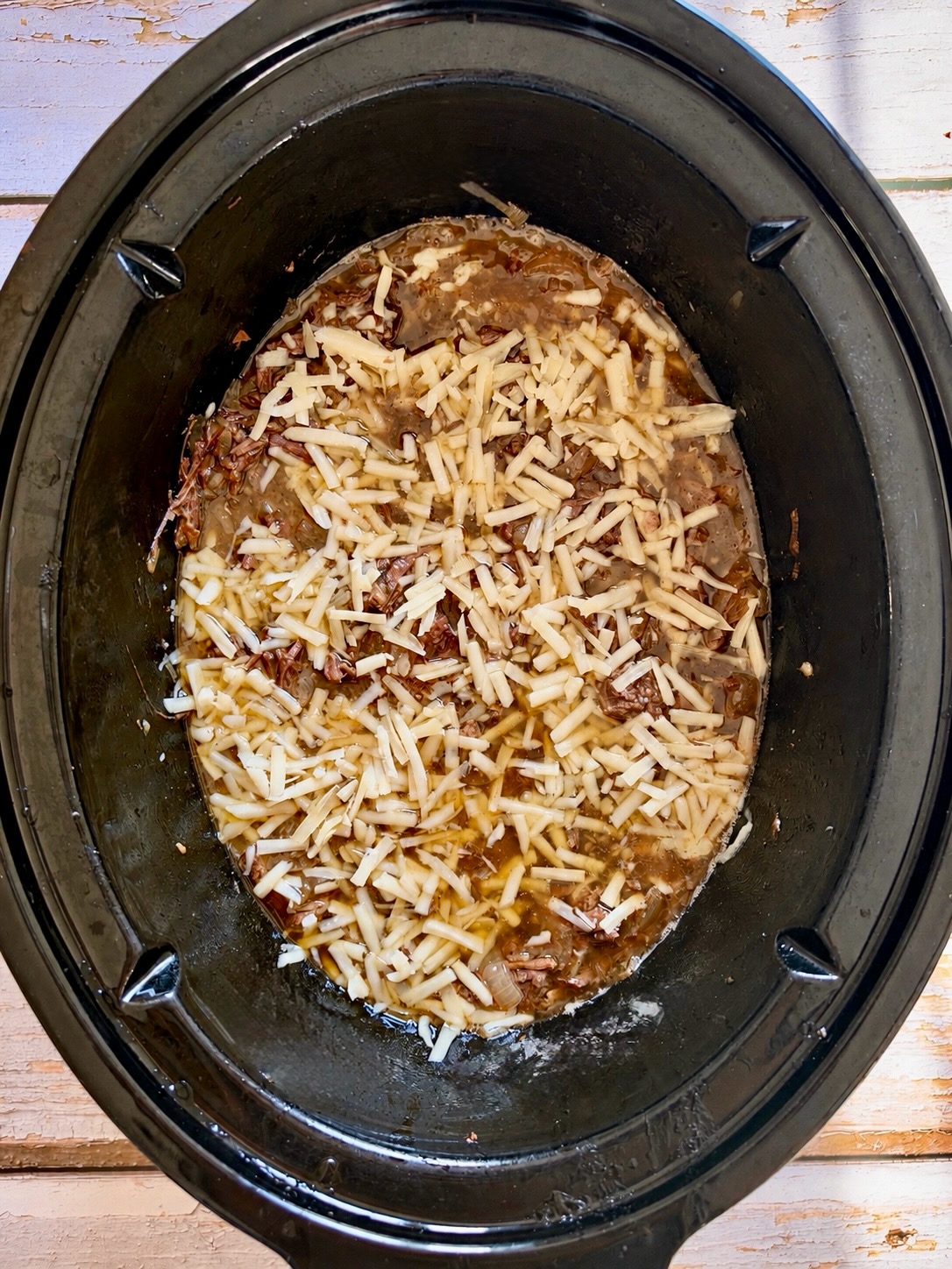 Shredded French onion pot roast topped with shredded Gruyère cheese in a slow cooker.