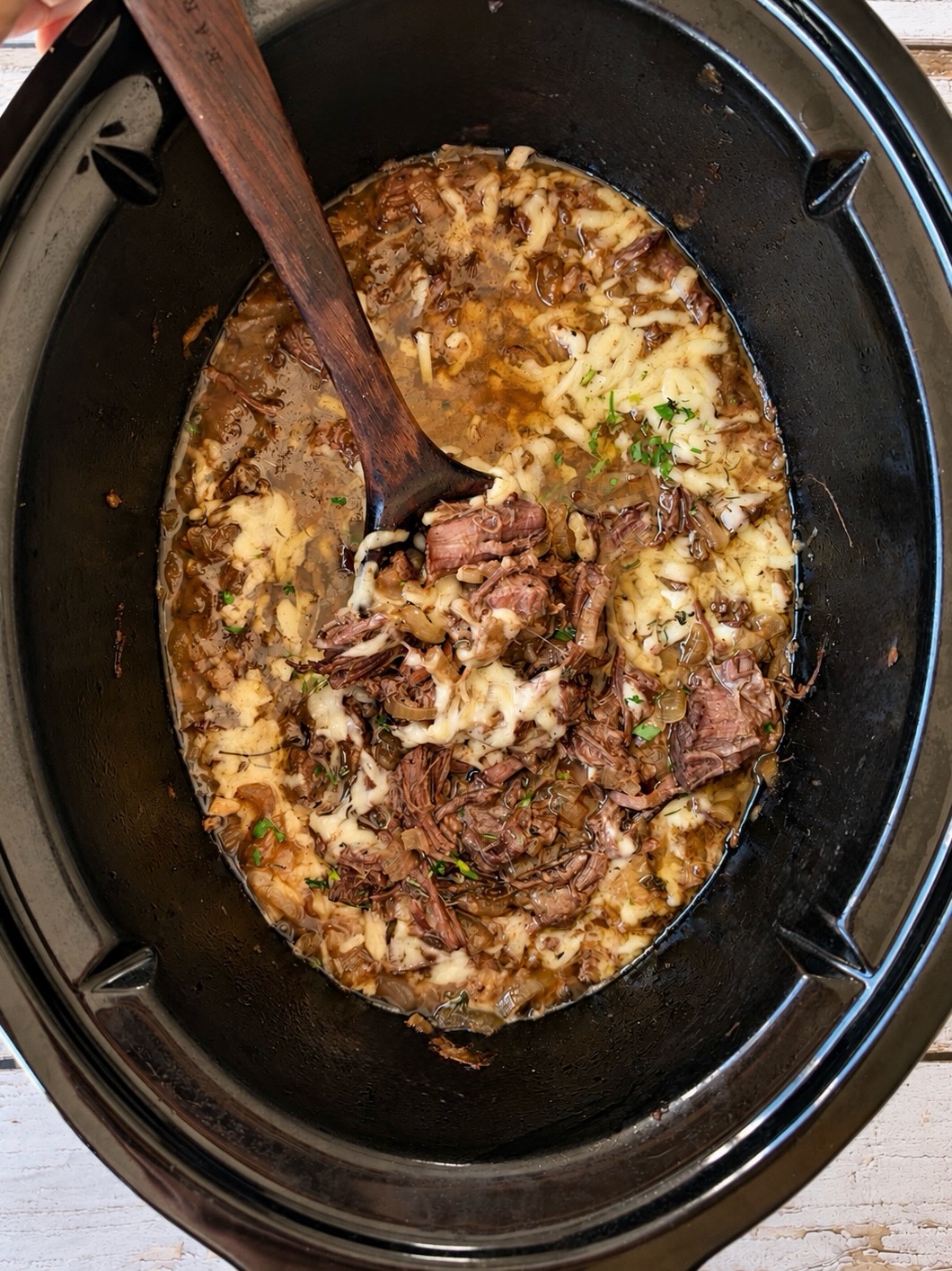 A wooden spoon stirring shredded beef with melted Gruyère and onions in a slow cooker.