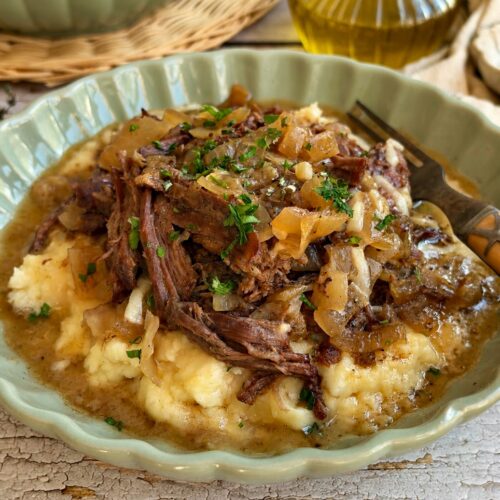 Crockpot French onion pot roast with shredded beef and caramelized onions served over mashed potatoes.