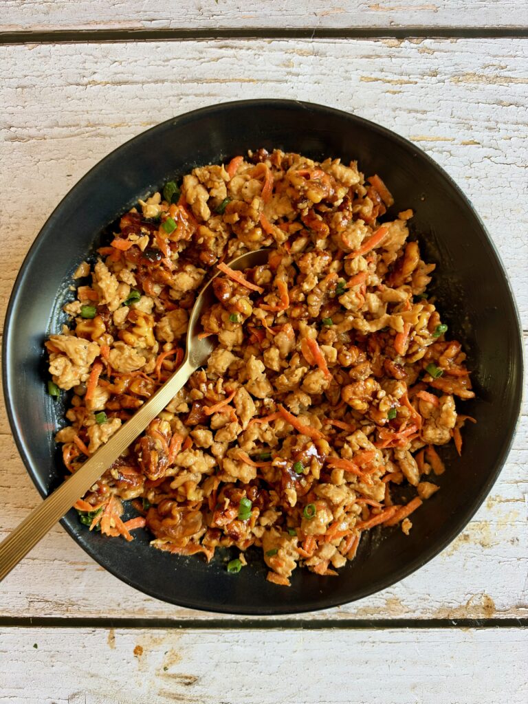 A black bowl filled with ground meat stir-fry with shredded carrots and green onions, served with a gold spoon.