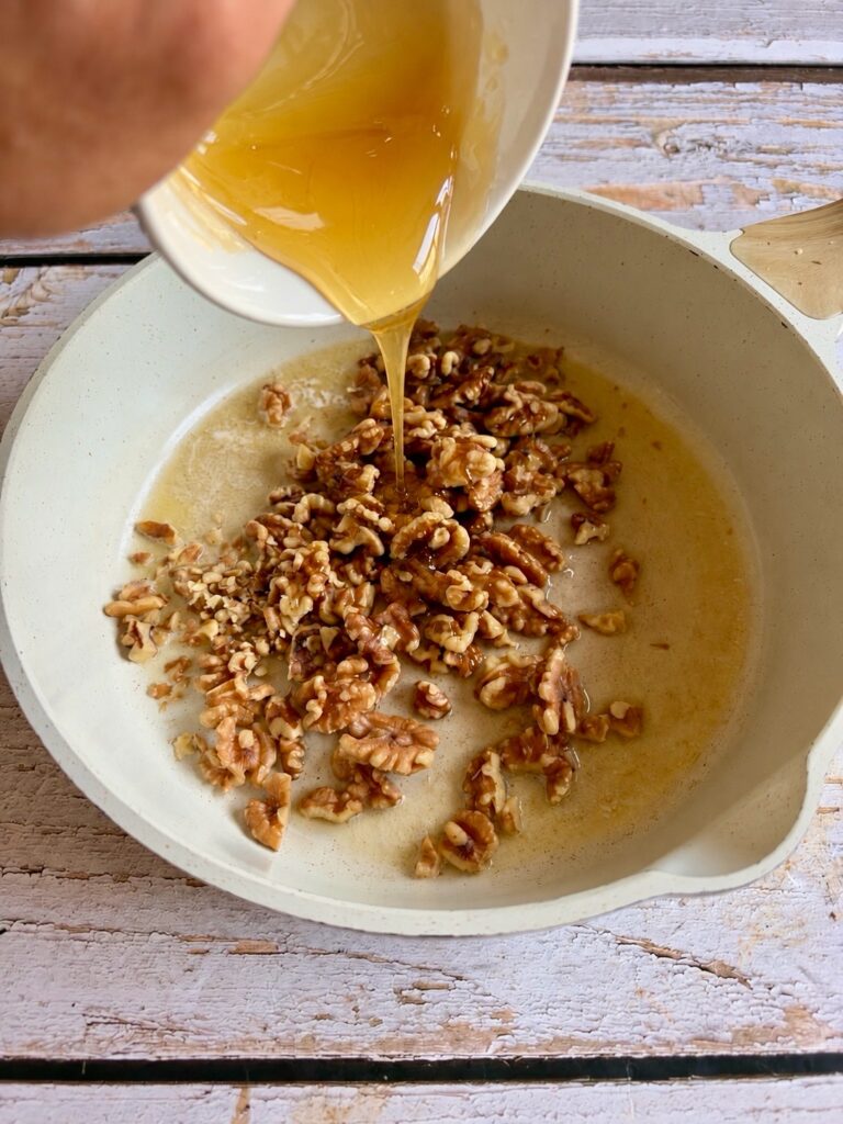 Honey being poured from a small bowl onto walnuts in a frying pan.