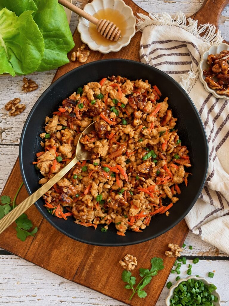 A black skillet filled with ground chicken and carrot stir-fry topped with green onions, resting on a wooden board with lettuce leaves, walnuts, and a honey dipper nearby.