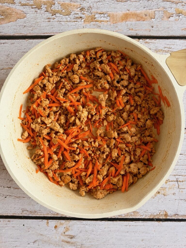 Ground meat and shredded carrots cooking in a light-colored nonstick pan.