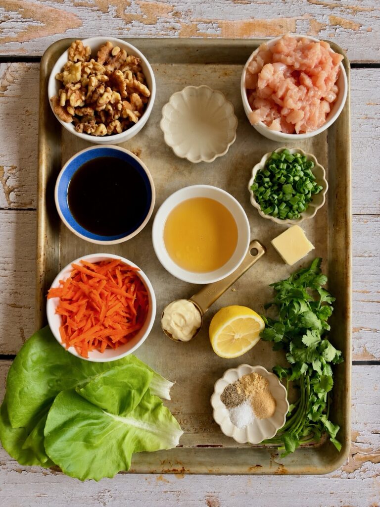 A baking sheet arranged with ingredients for a chicken salad recipe, including chopped chicken, walnuts, carrots, lettuce, herbs, spices, and liquid condiments.