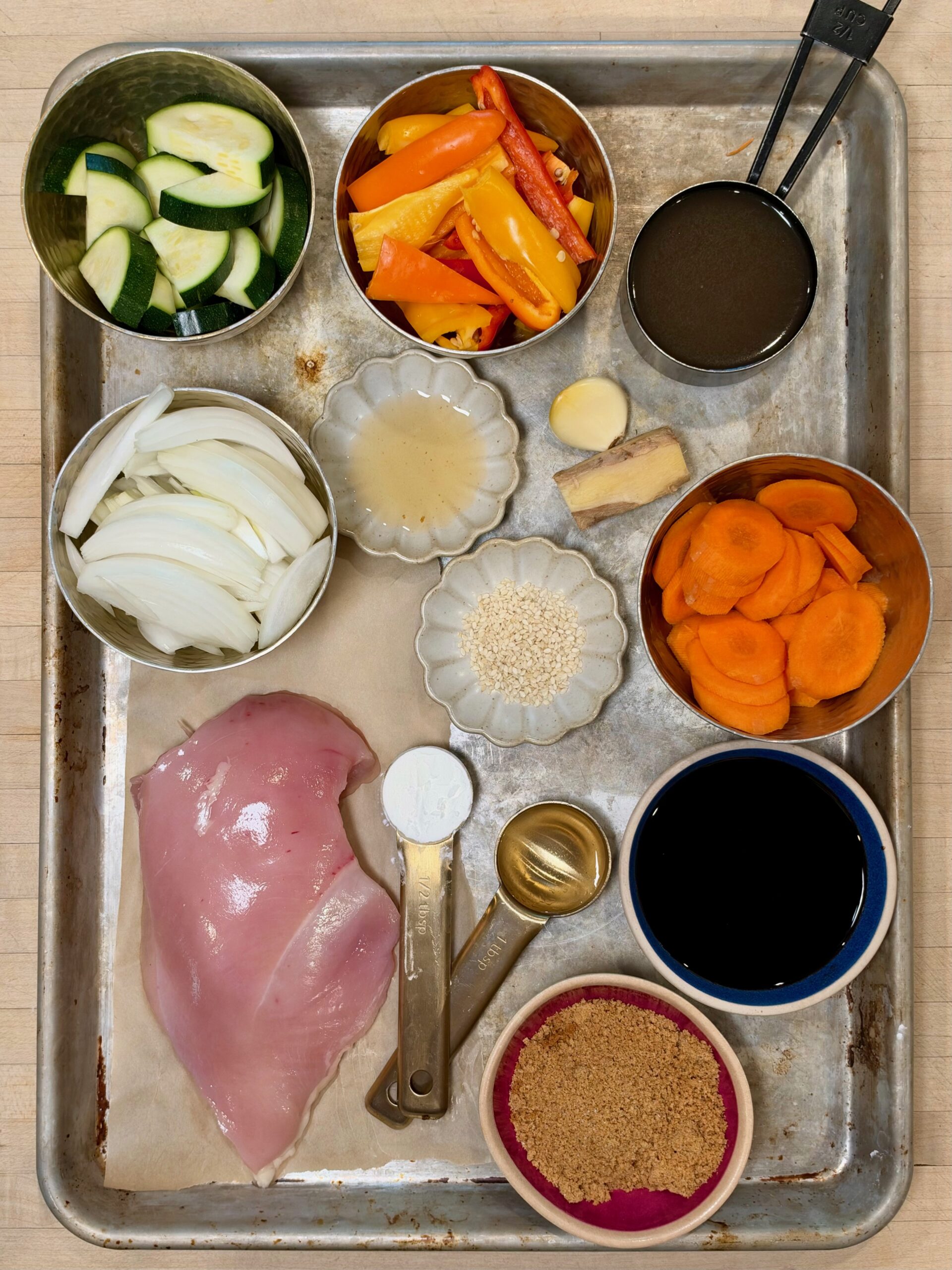 A metal sheet pan arranged with ingredients for a meal, including sliced chicken breast, zucchini, bell peppers, carrots, onions, ginger, garlic, sesame seeds, brown sugar, soy sauce, and measured liquid and dry seasonings.