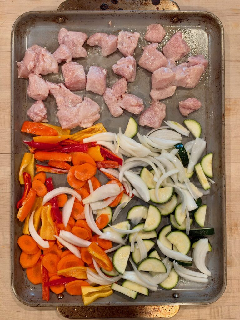 A baking sheet containing raw diced chicken, sliced orange bell peppers, carrots, zucchini, and onions.