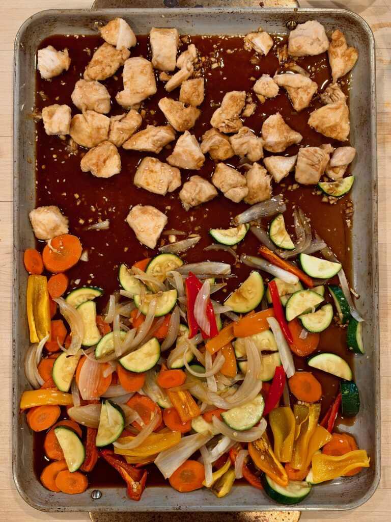 A sheet pan containing cooked chicken chunks in a dark sauce on one side and a mix of roasted sliced zucchini, carrots, onions, and bell peppers on the other.