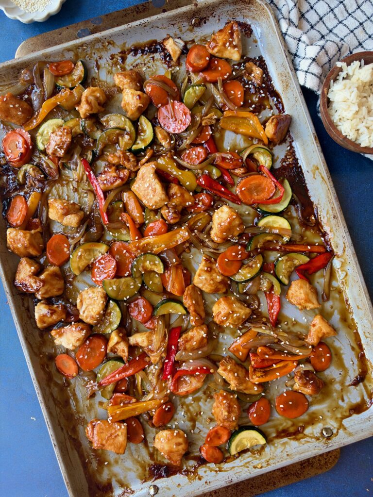 A sheet pan meal of stir-fried chicken, sliced carrots, zucchini, and bell peppers coated in a dark glaze and sprinkled with sesame seeds.