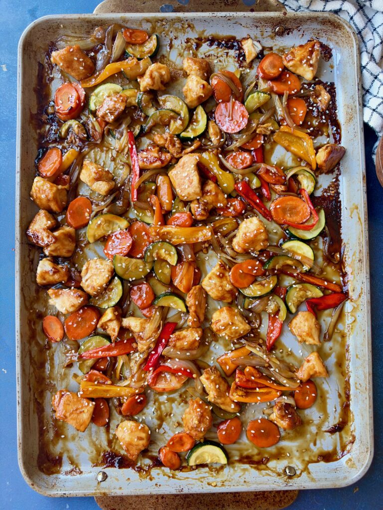 A sheet pan filled with roasted chicken, sliced zucchini, carrots, and bell peppers in a dark glaze, sprinkled with sesame seeds.