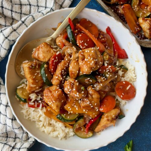 A bowl of chicken and vegetable stir-fry served over white rice, topped with sesame seeds and a gold serving spoon.