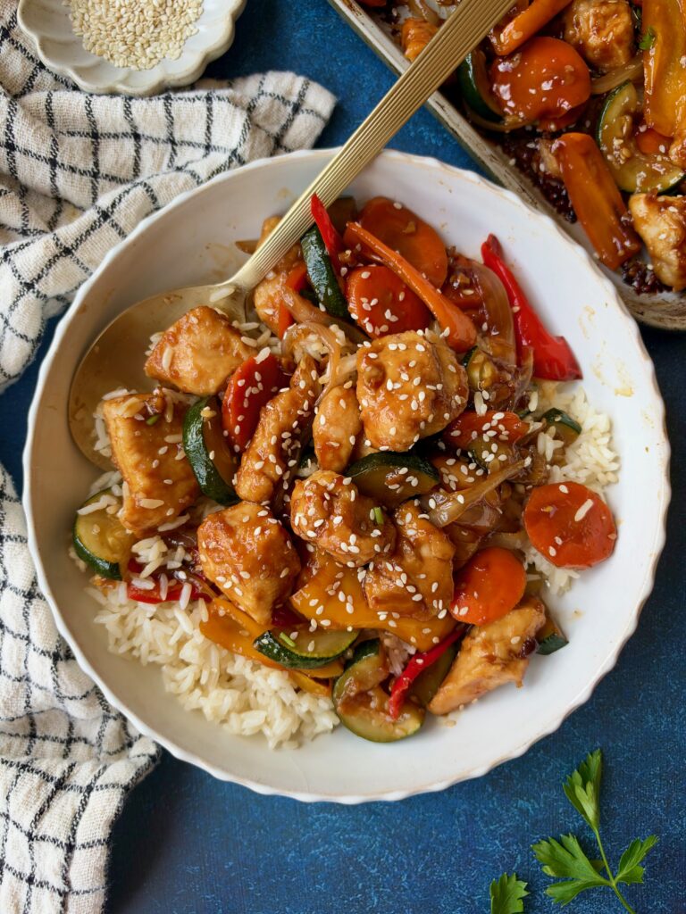 A bowl of chicken and vegetable stir-fry served over white rice, topped with sesame seeds and a gold serving spoon.