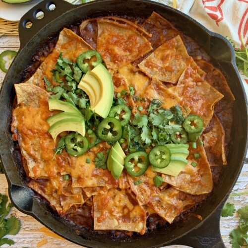 Skillet beef enchiladas topped with melted cheese, avocado, jalapeño, and cilantro in a cast iron skillet.