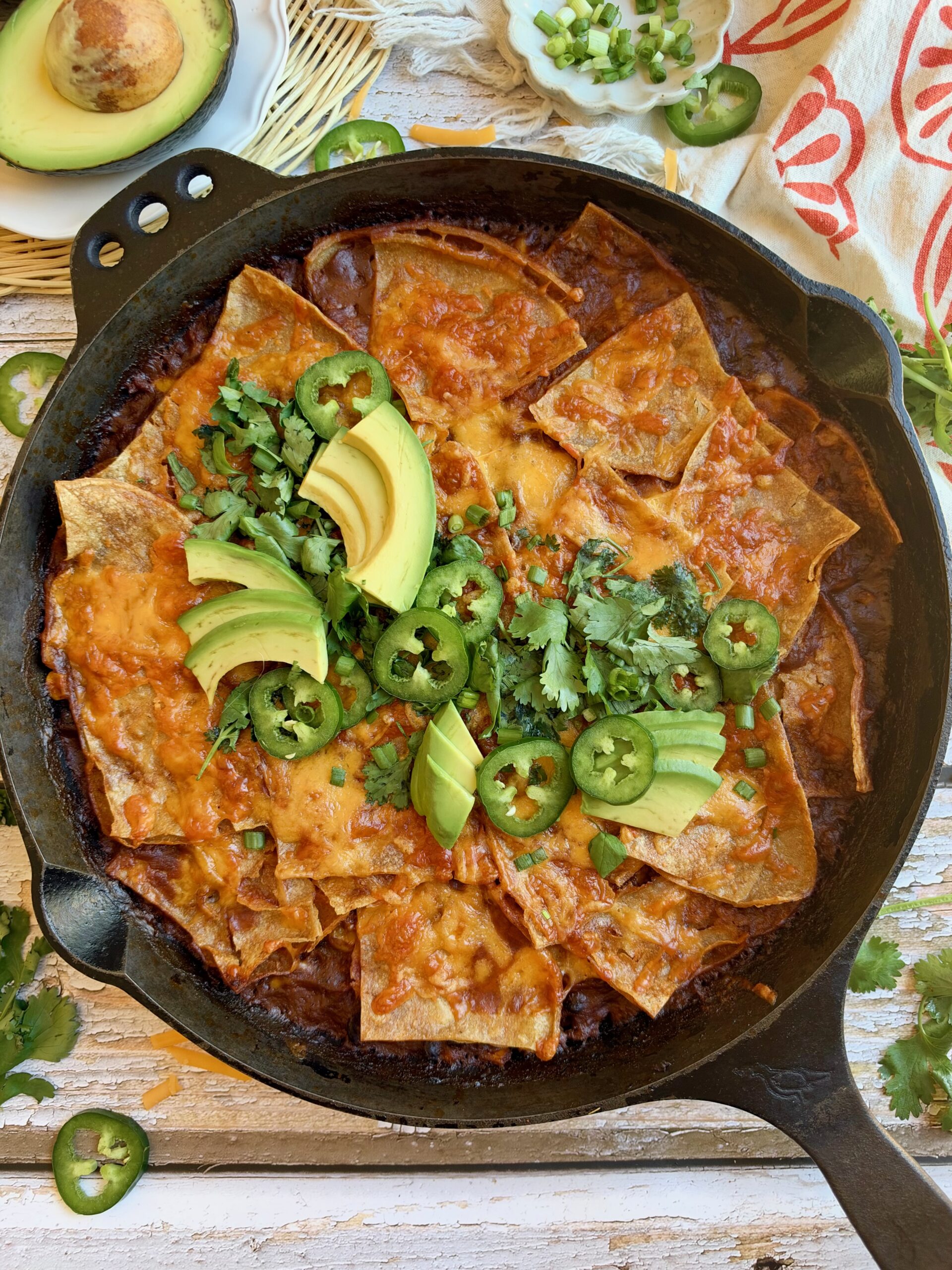 Skillet beef enchiladas topped with melted cheese, avocado, jalapeño, and cilantro in a cast iron skillet.