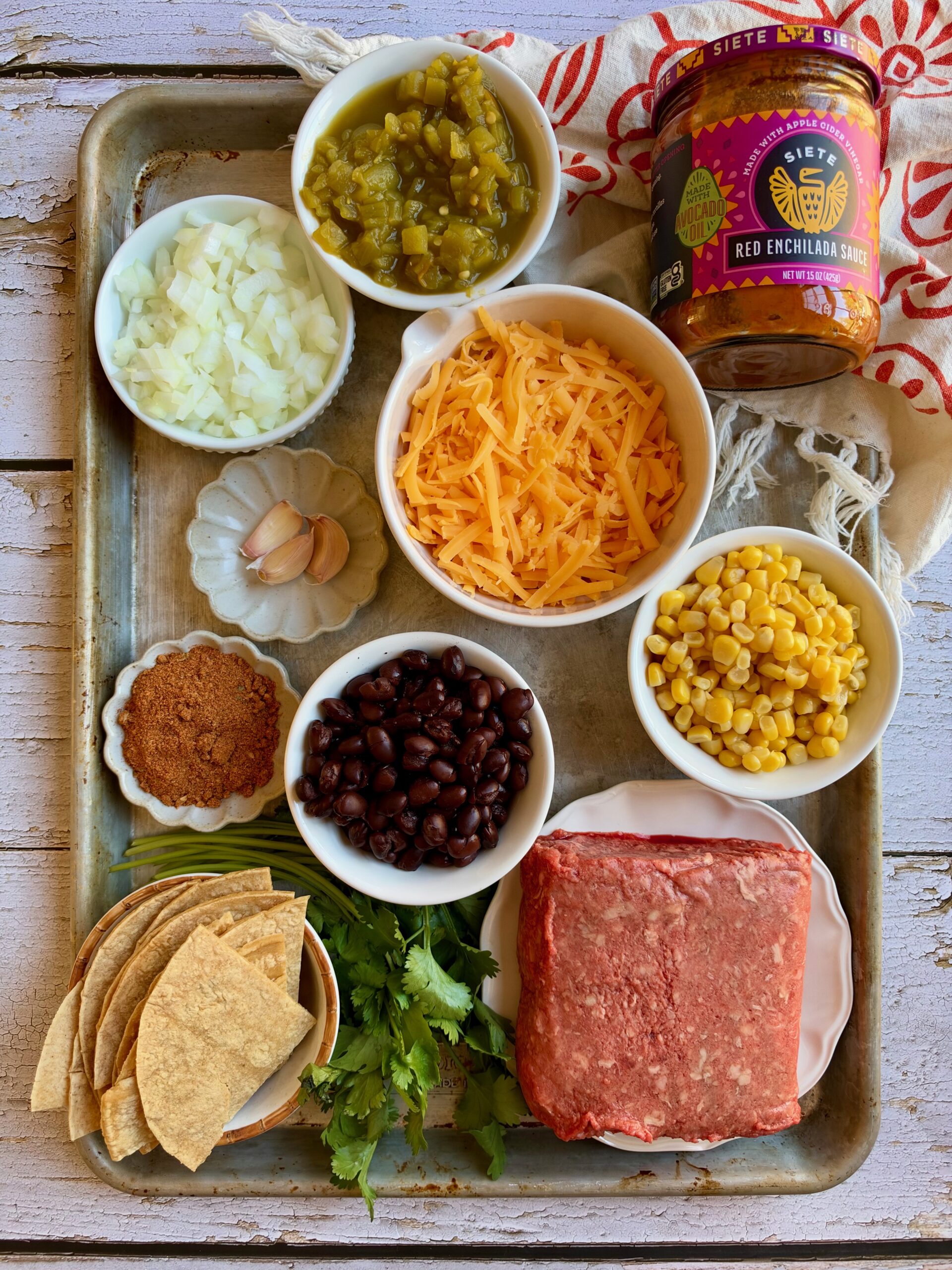 Ingredients for skillet beef enchiladas on a tray, including ground beef, enchilada sauce, black beans, corn, green chiles, shredded cheese, taco seasoning, onion, garlic, and corn tortillas.