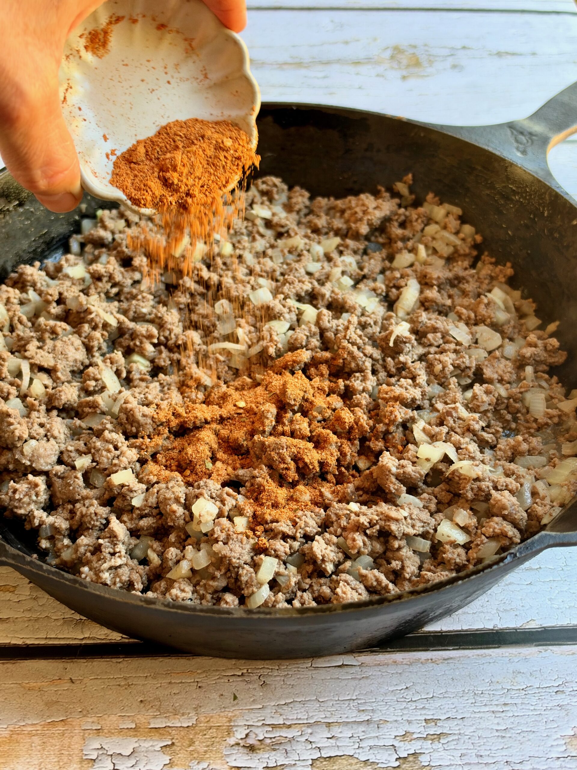 Taco seasoning being sprinkled over browned ground beef in a cast iron skillet.