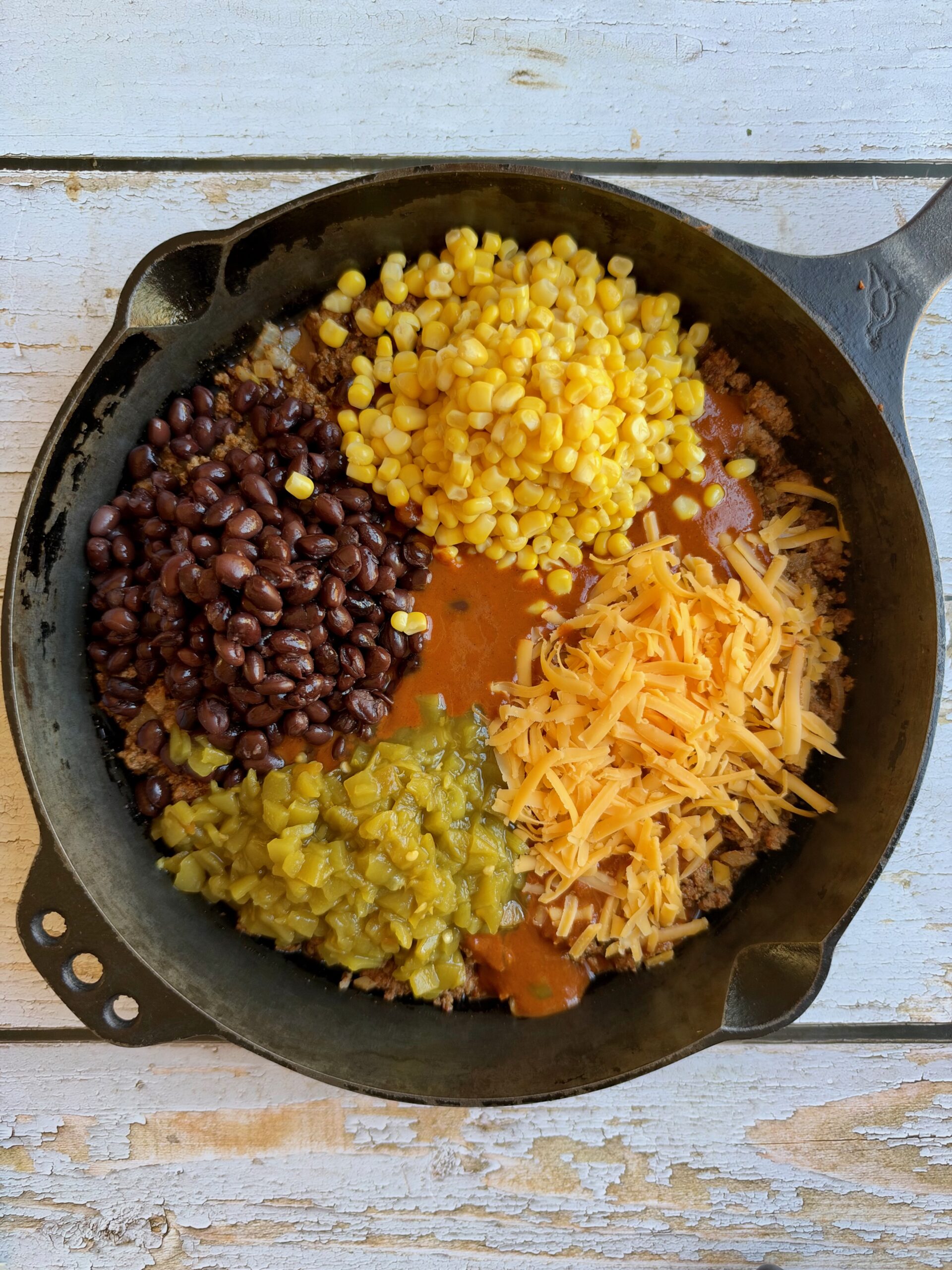 Black beans, corn, green chiles, and shredded cheese added on top of seasoned ground beef in a cast iron skillet.