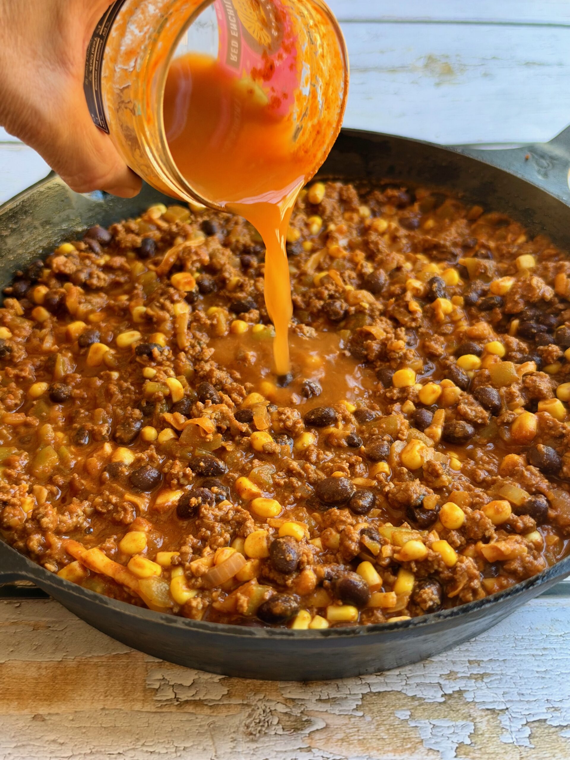 Red enchilada sauce being poured over ground beef, black beans, and corn in a cast iron skillet.