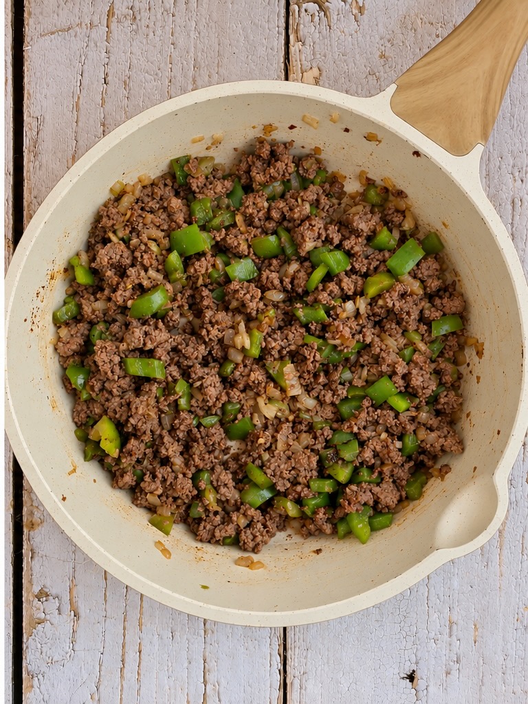 Ground beef sautéed with diced green bell peppers and onions in a light-colored skillet.