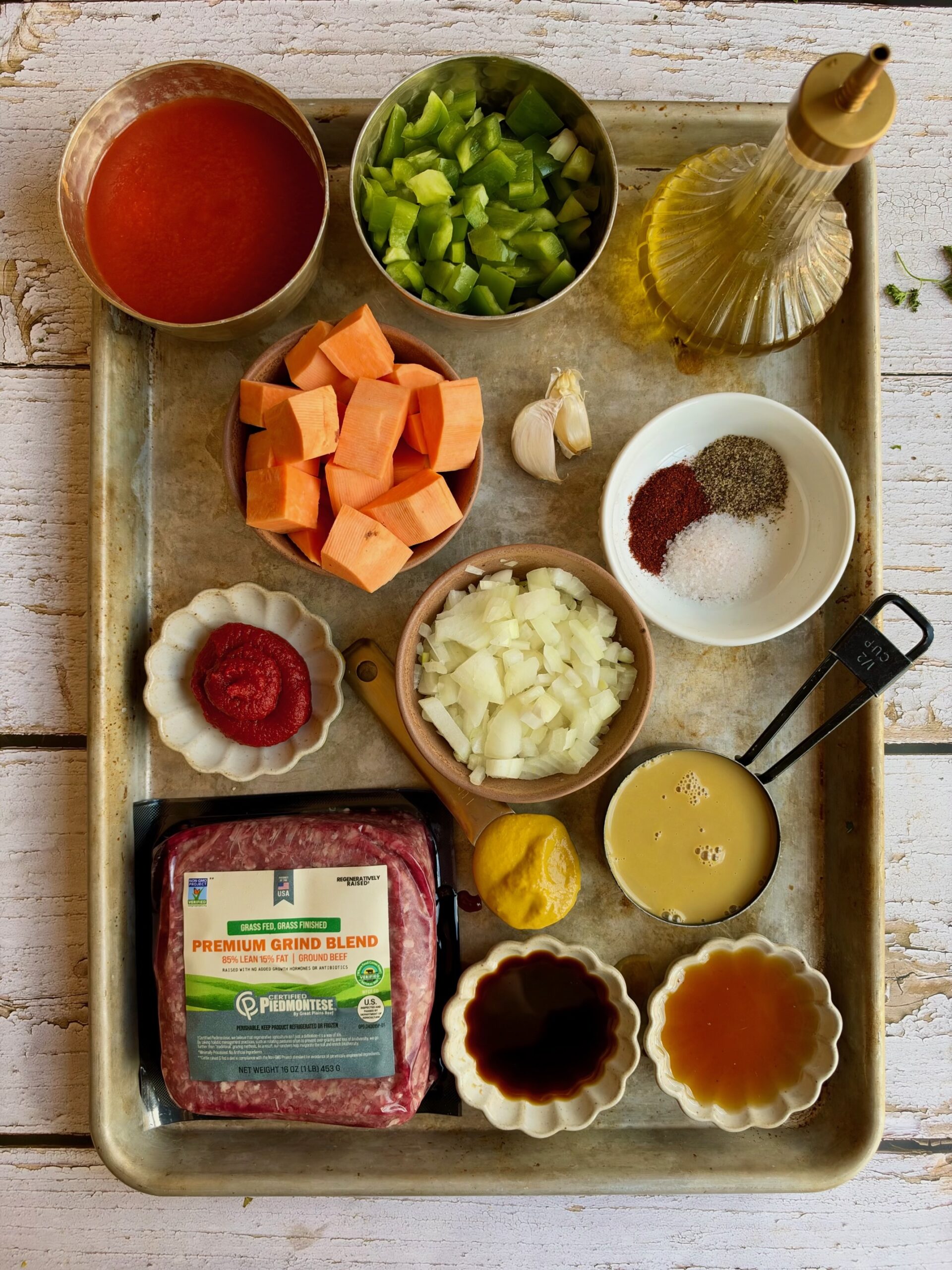 A sheet pan filled with ingredients for a meal, including a package of ground beef, diced sweet potatoes, chopped green peppers and onions, tomato paste, garlic cloves, broth, mustard, oil, and various spices and liquids in small bowls.