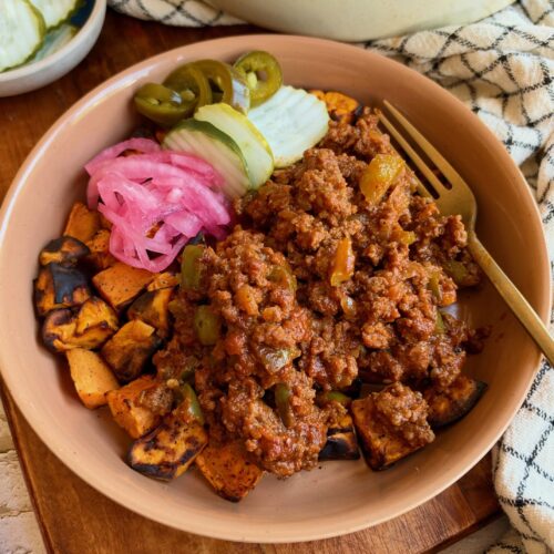 A bowl filled with seasoned ground meat over roasted sweet potato cubes, topped with pickled red onions, sliced jalapeños, and cucumber slices, served with a fork.