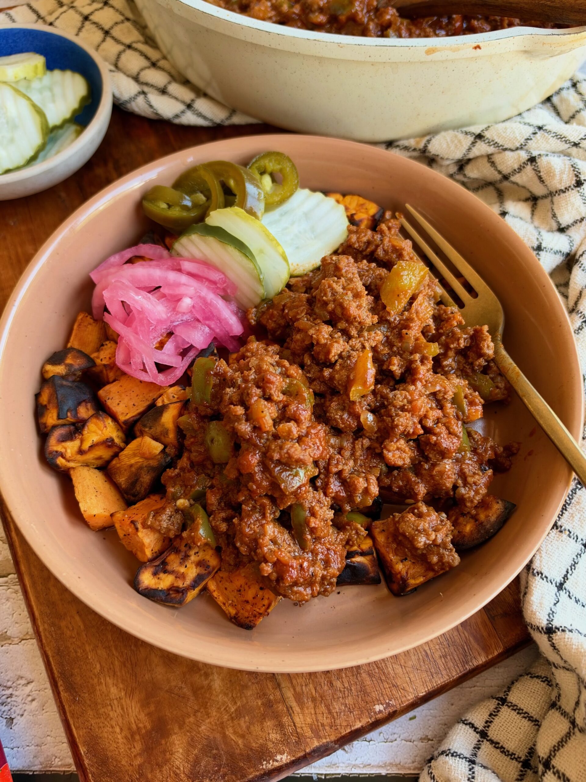 A bowl filled with seasoned ground meat over roasted sweet potato cubes, topped with pickled red onions, sliced jalapeños, and cucumber slices, served with a fork.