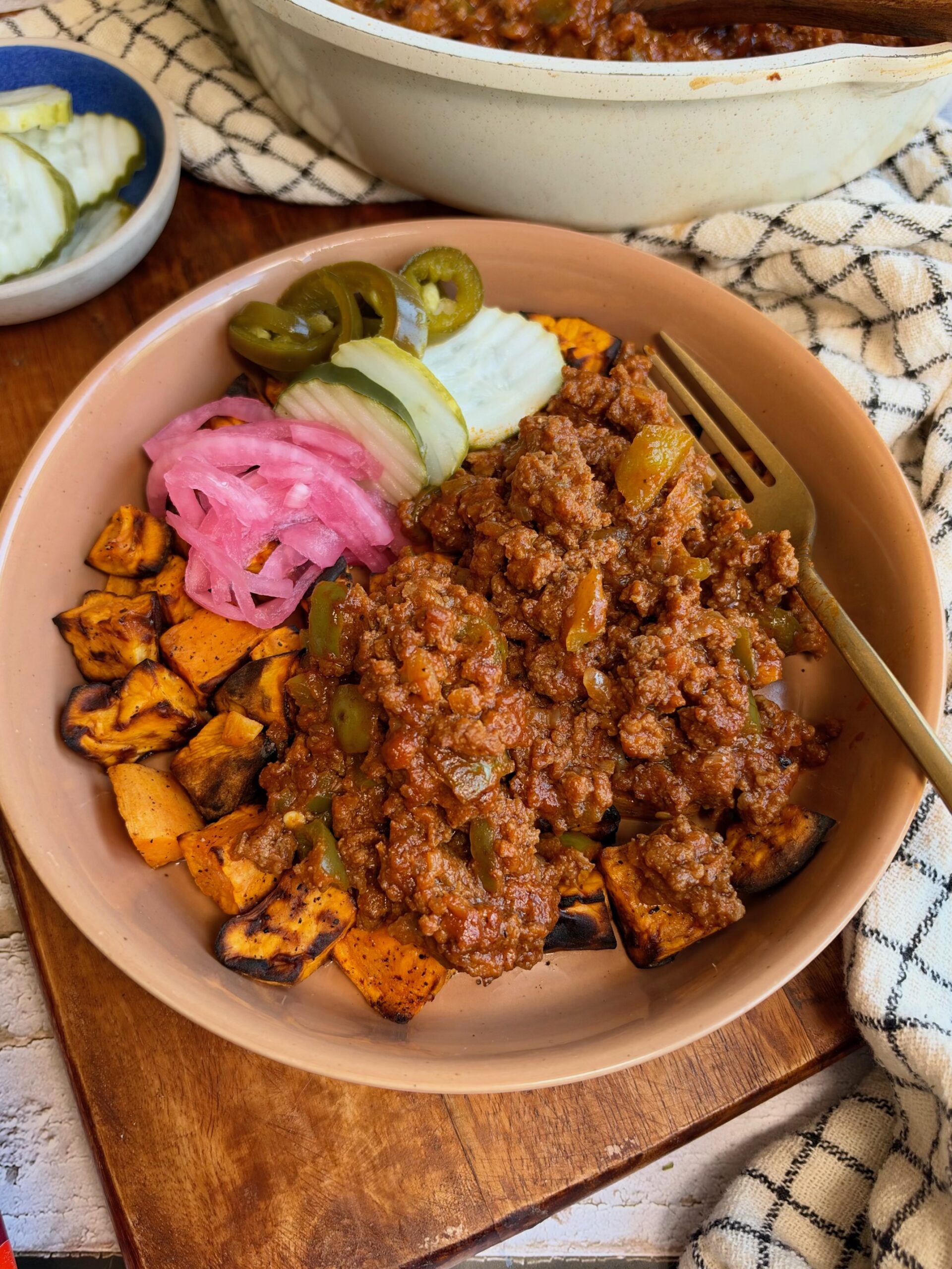 A bowl of ground meat sauce over roasted sweet potato cubes, topped with pickled red onions, sliced cucumbers, and jalapeños, with a fork resting on the side.