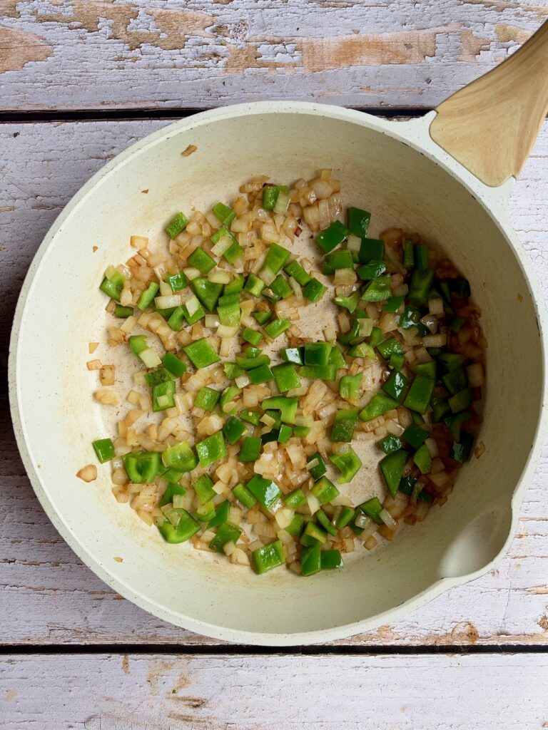 Chopped green bell peppers and onions sautéing in a light-colored pan.