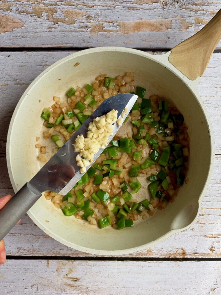 A knife blade holding minced garlic over a pan of sautéed onions and green peppers.