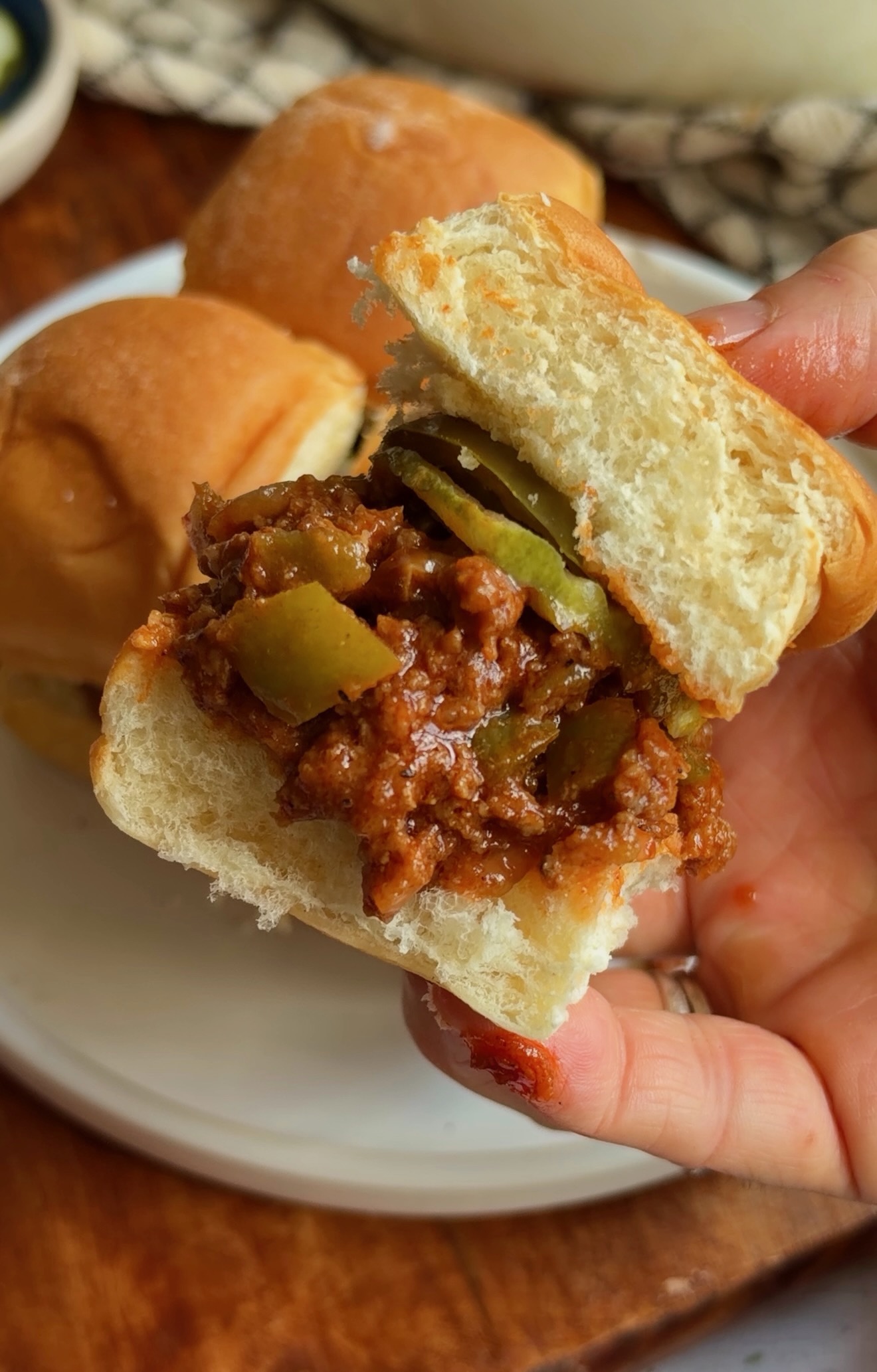 A hand holding a slider filled with sloppy joe meat and sliced pickles, with more sliders on a plate in the background.