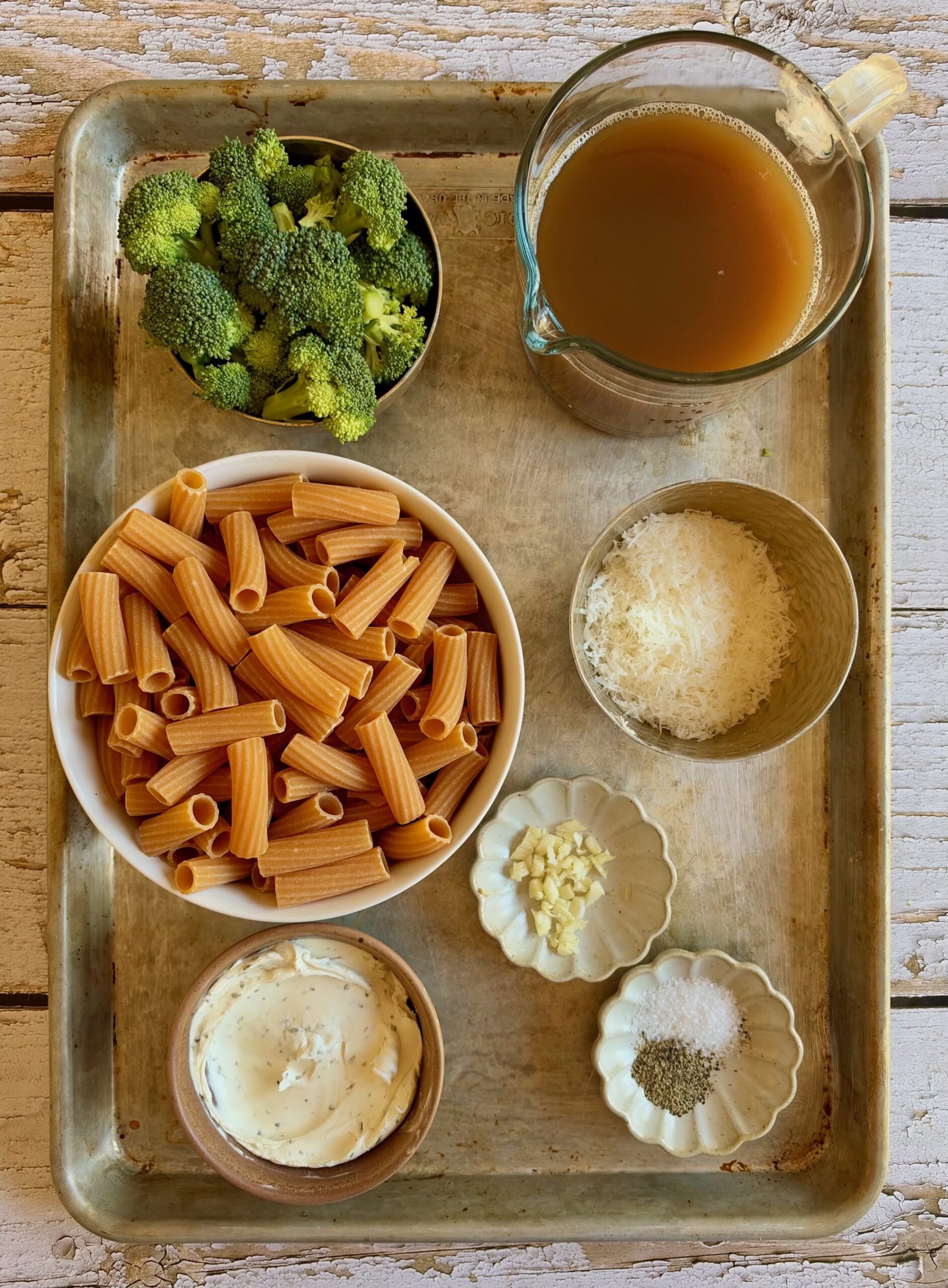 A metal tray containing ingredients for pasta: rigatoni, broccoli florets, a pitcher of broth, shredded cheese, cream cheese, minced garlic, salt, and pepper.