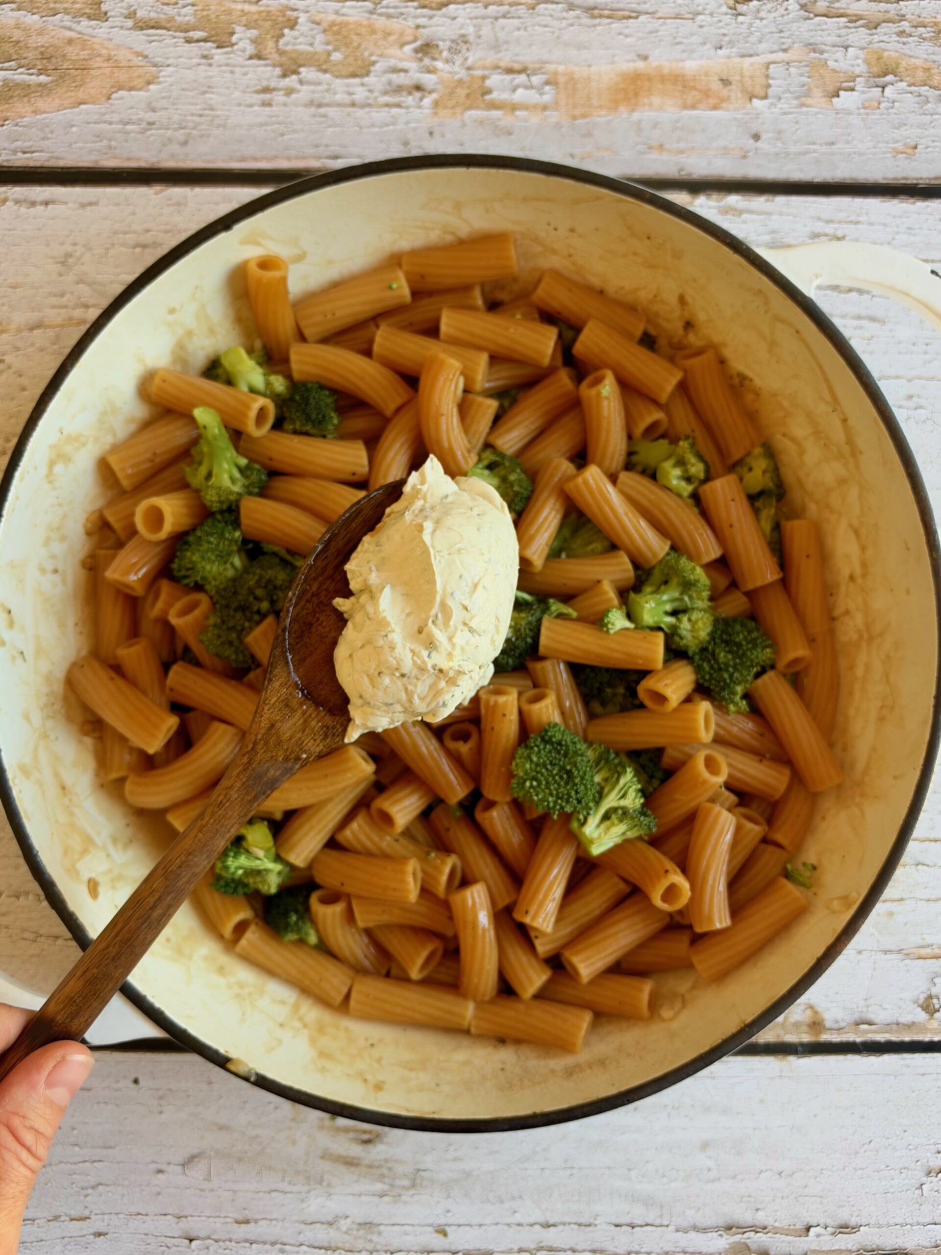 A wooden spoon holding a scoop of herbed cream cheese over a pan of rigatoni pasta and broccoli florets.
