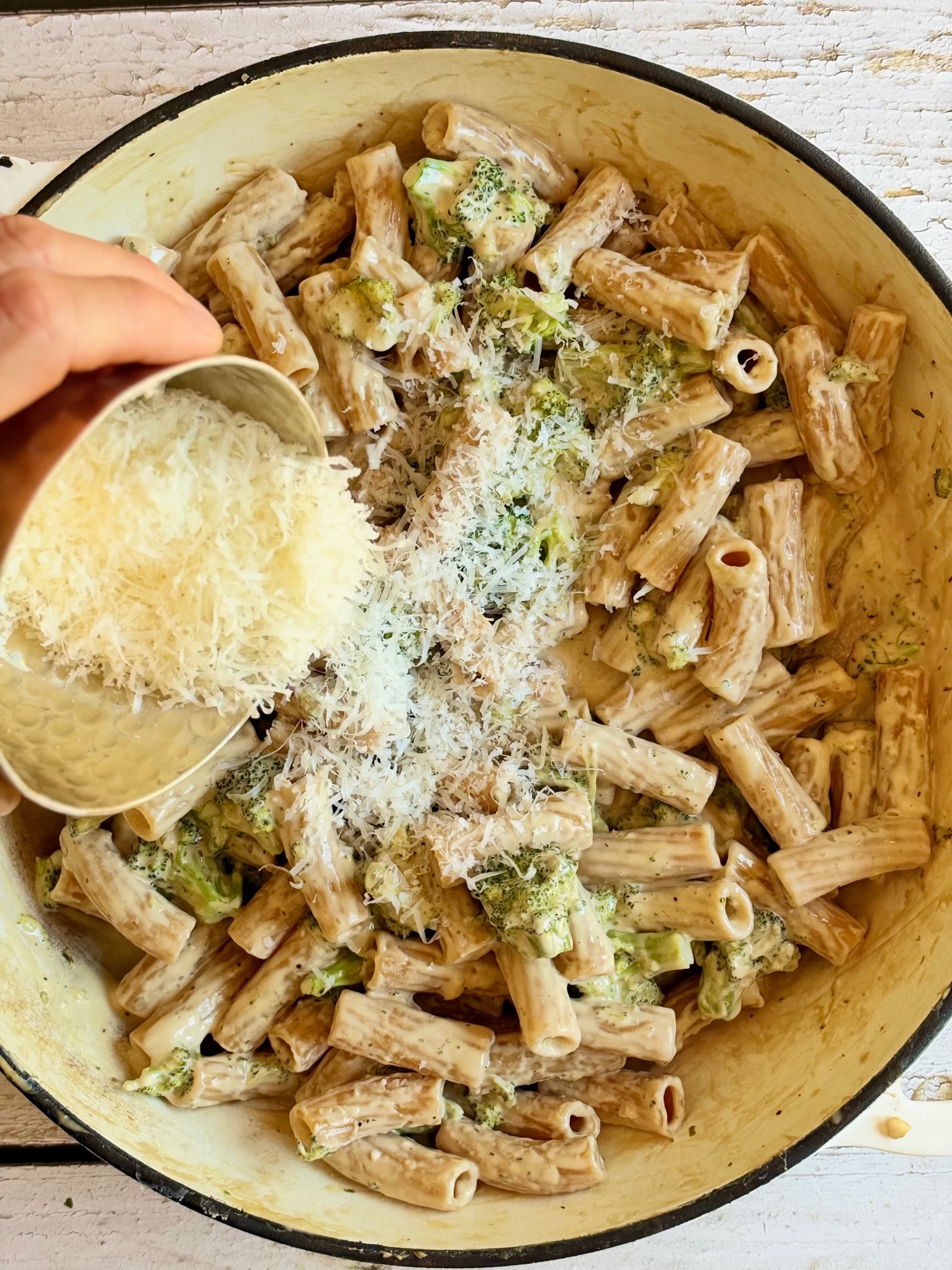 A hand sprinkles grated parmesan cheese over a pot of creamy pasta with broccoli.