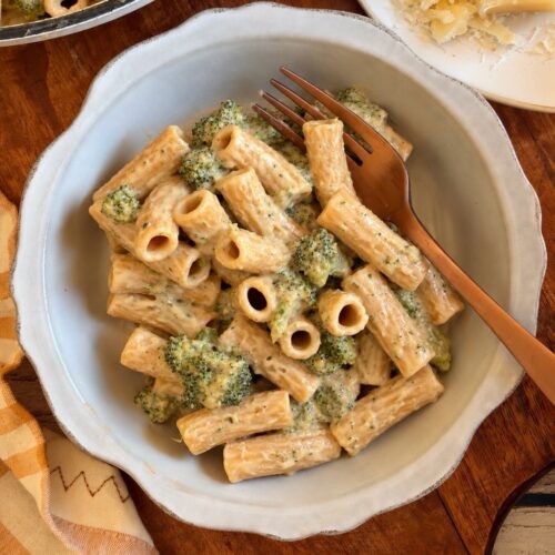 Rigatoni pasta with broccoli in a creamy sauce in a bowl with a fork, next to a block of parmesan cheese.